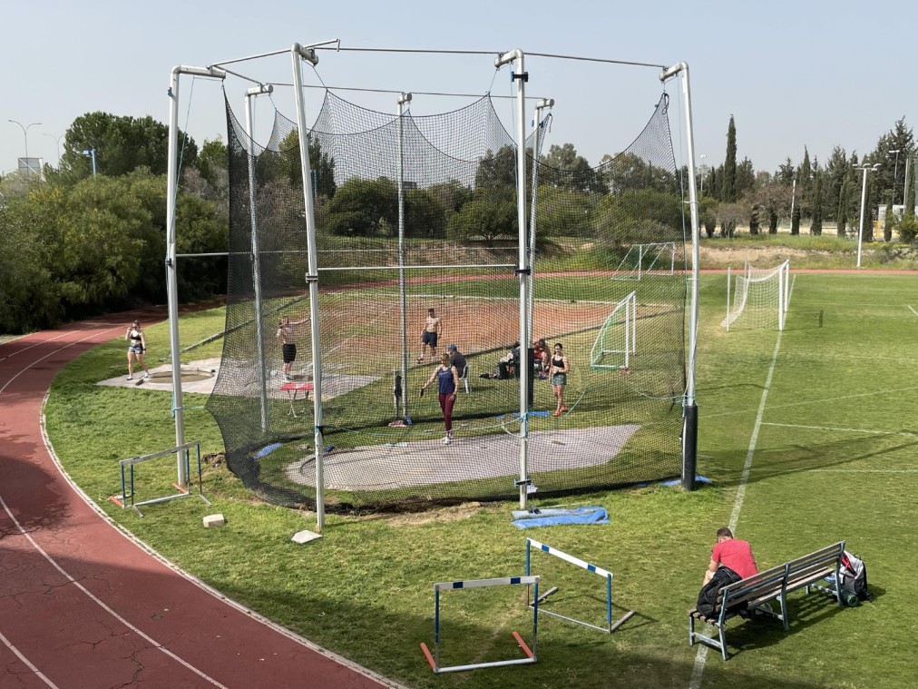 Athletes practicing discus or hammer throw inside netted cage on grassy training field