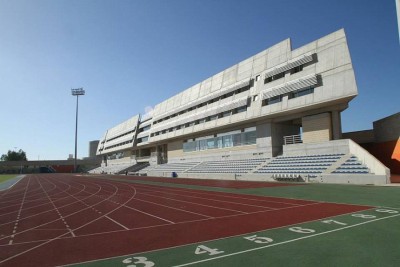 Modern athletics stadium with red running track and large grandstand under clear blue sky