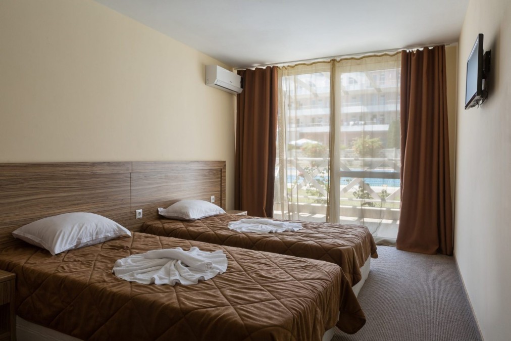Twin-bed hotel room with brown bedding and balcony view, lodging for athletes during training camp