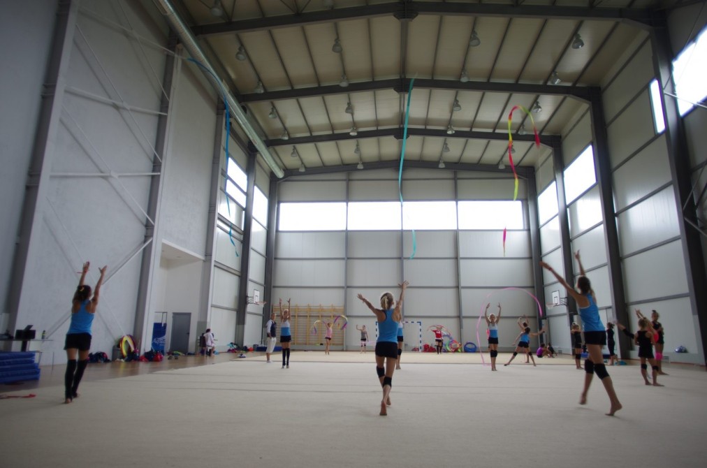 Athletes practicing rhythmic gymnastics with ribbons in a large indoor hall during a training camp session