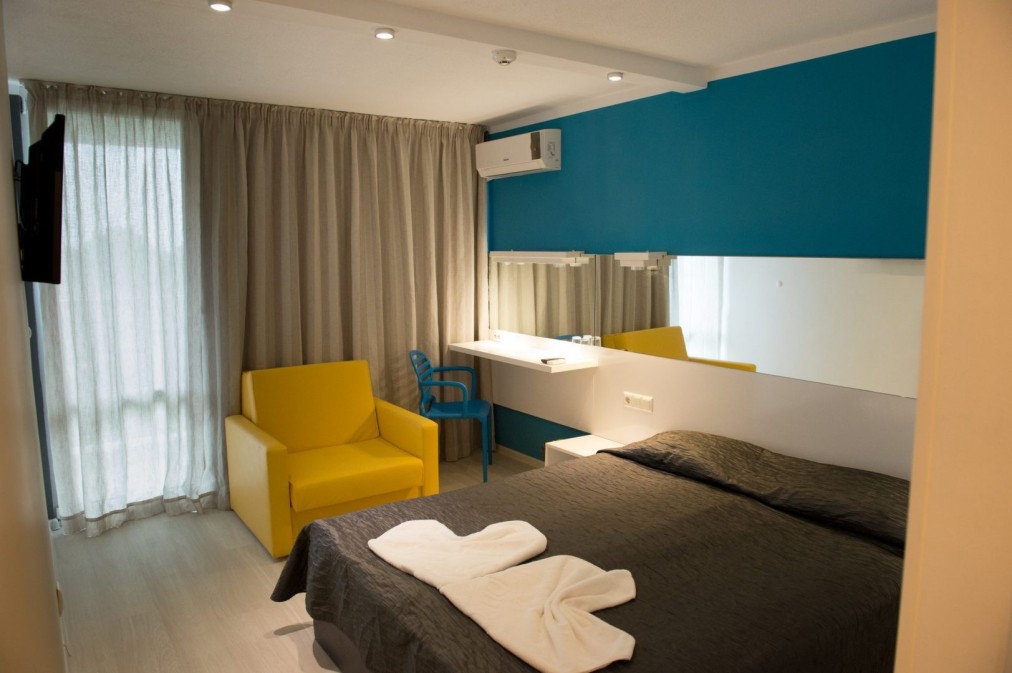 Modern hotel room with dark bedspread, yellow chair, and desk, athlete lodging during training camp