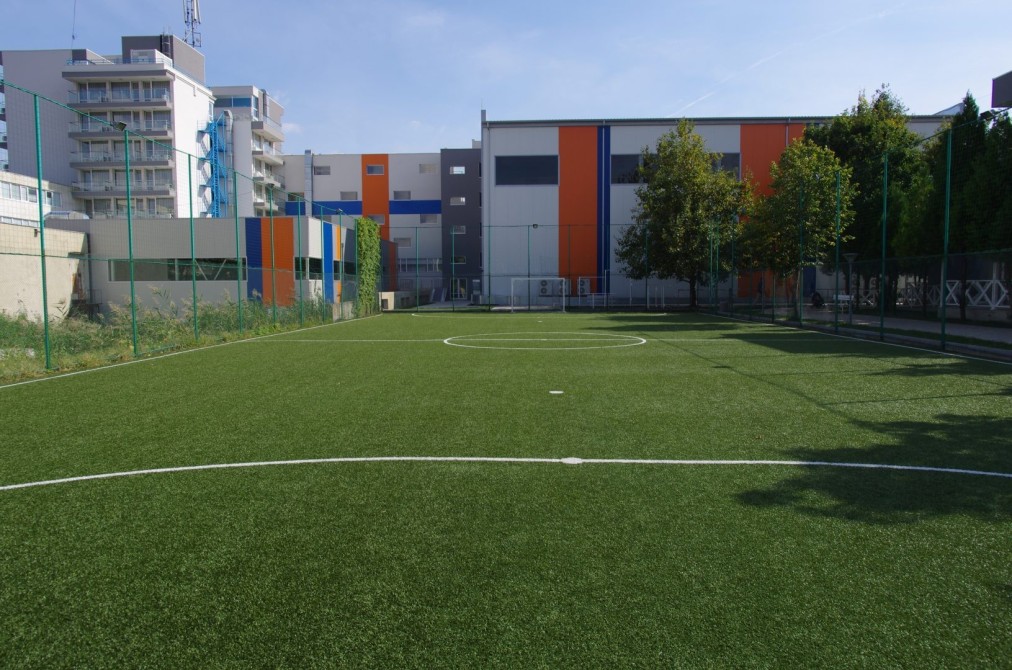 Green mini soccer pitch with white lines, surrounded by campus buildings for training camp drills