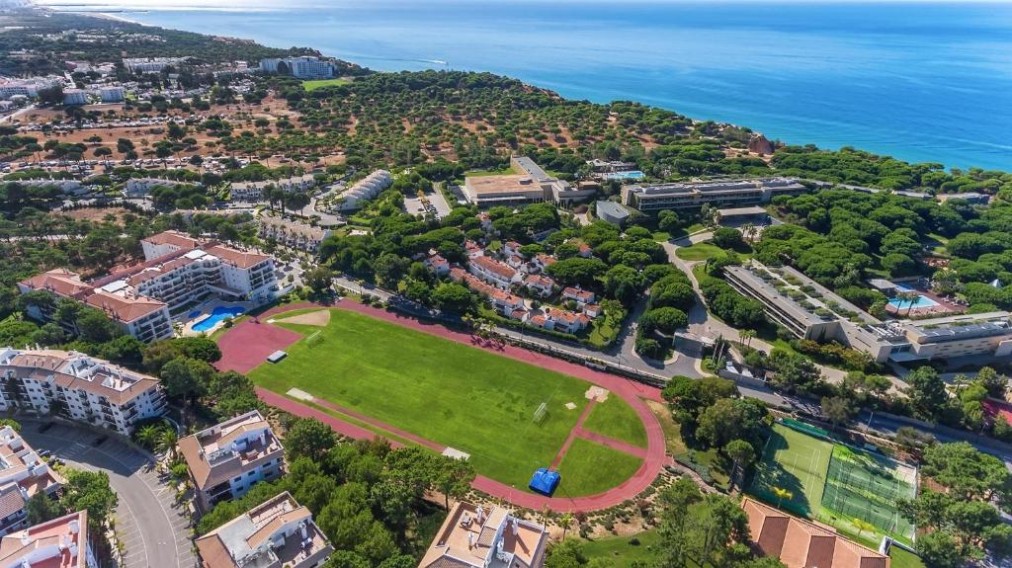 Aerial overview of a coastal sports complex with athletics track, fields, and courts.