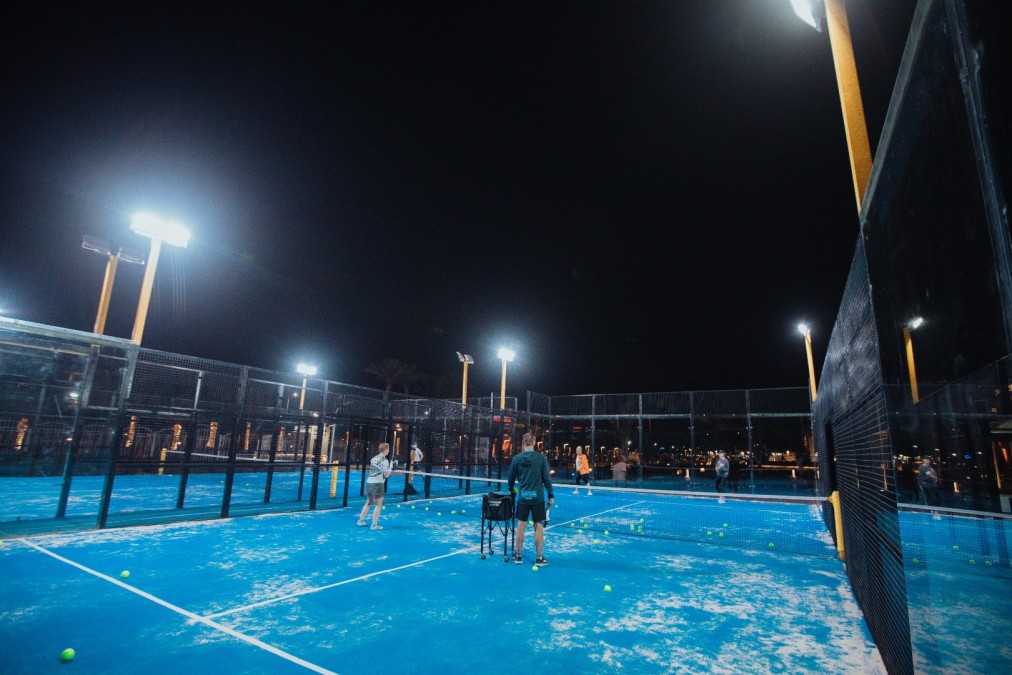 Night padel session on blue outdoor courts under floodlights, with a ball basket at the net.