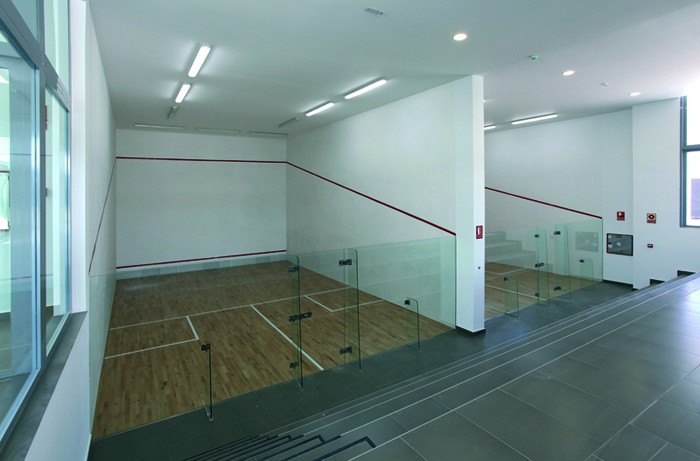 Indoor squash court with glass back wall, wooden floor, and overhead lighting.
