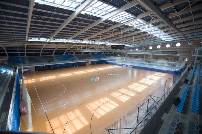 Large indoor sports hall with marked court, tiered seating, and skylights.