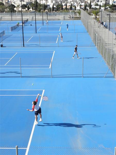 Outdoor tennis courts with blue surface and fencing, players practicing on several courts.