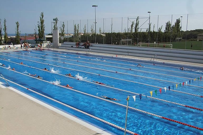 Athletes swim laps in an outdoor lane pool beside sports fields.