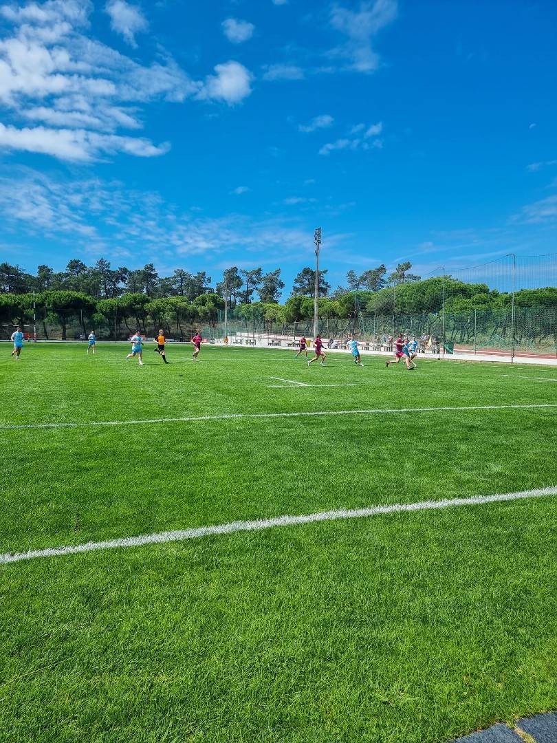 Football match in progress on a grass field under a bright blue sky.