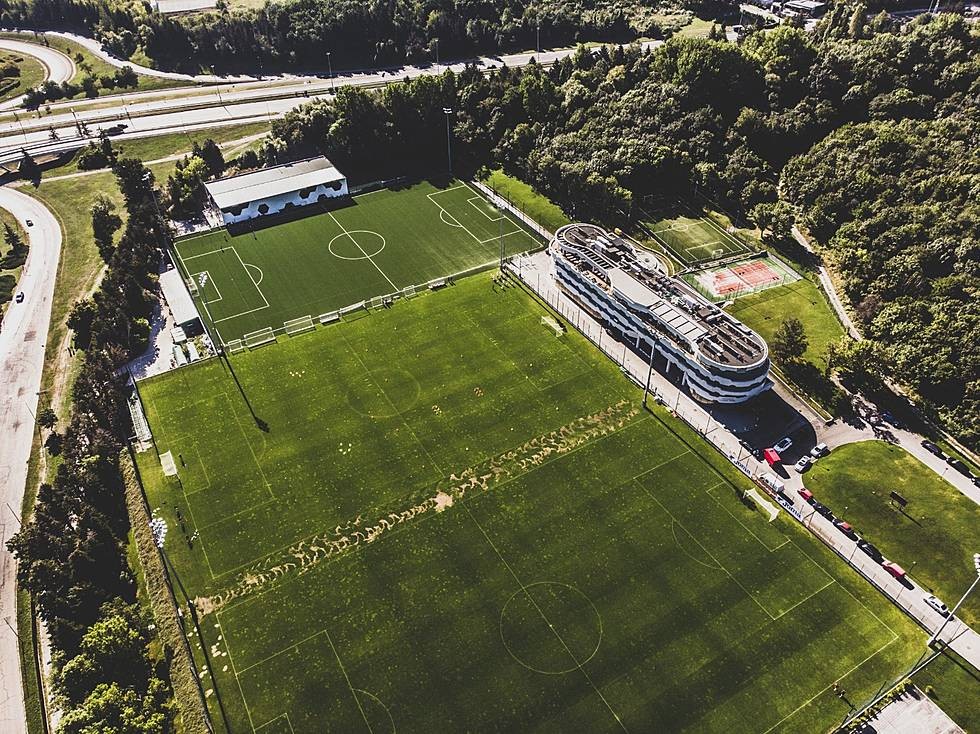 Aerial view of multiple green soccer fields beside a modern building, set in forested hills for training camp practice