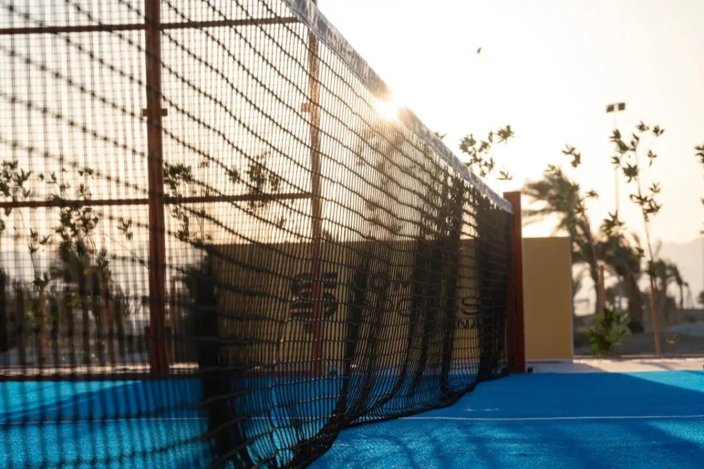 Close-up of a padel net on an outdoor blue court with low sun and palm trees in the background.