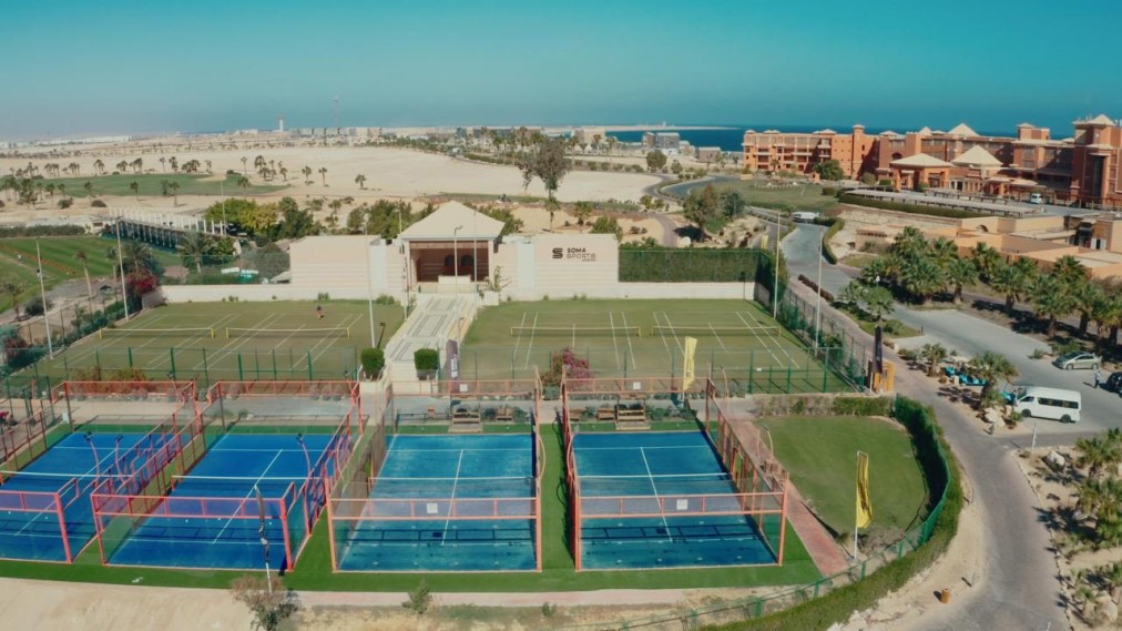 Aerial view of padel and tennis courts at a coastal sports complex with nearby resort buildings.