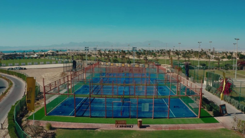 Wide view of an outdoor padel court complex with multiple blue courts and surrounding grounds.