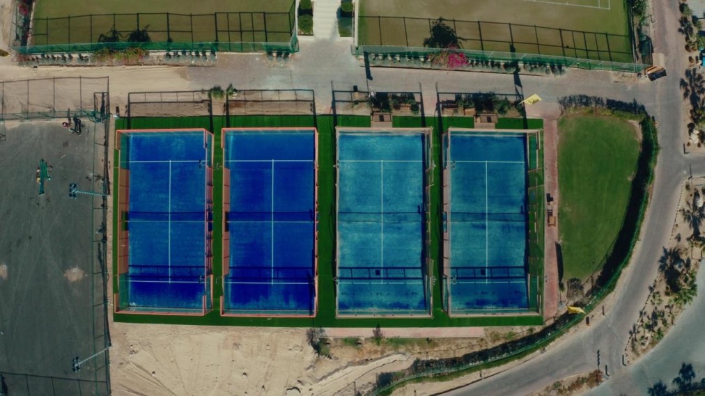 Aerial view of four outdoor padel courts with blue surfaces inside fenced enclosures.