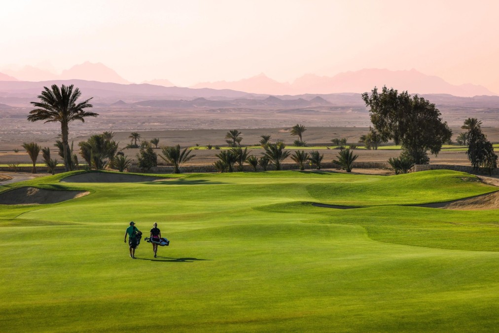 Two golfers carry bags across a fairway at dusk, palm trees and distant mountains under a pink sky