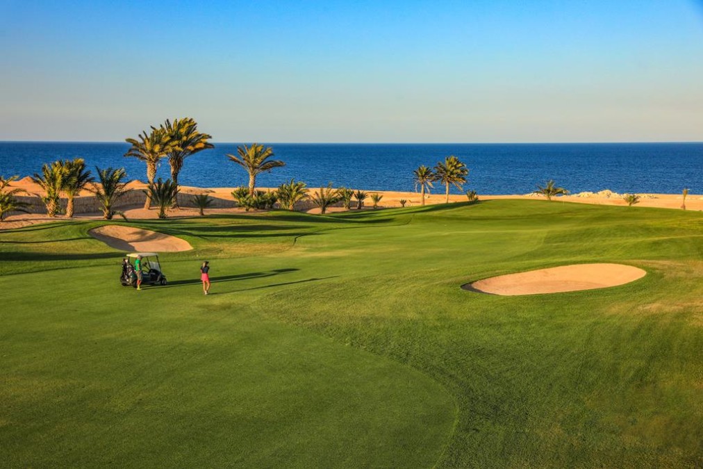 Golfer hits a shot near a cart on a coastal fairway, palm trees and sand bunkers by the sea