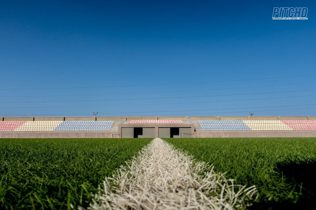 Low-angle view across football pitch showing stadium seating with multicolored stands