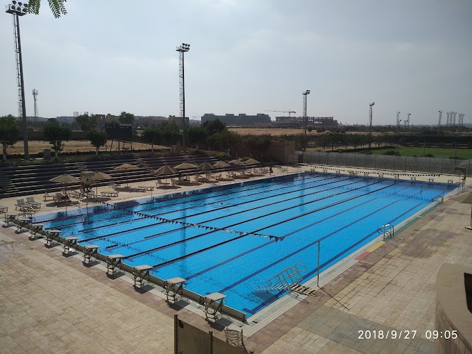 Outdoor competition swimming pool with lane ropes and starting blocks, surrounded by seating and umbrellas.