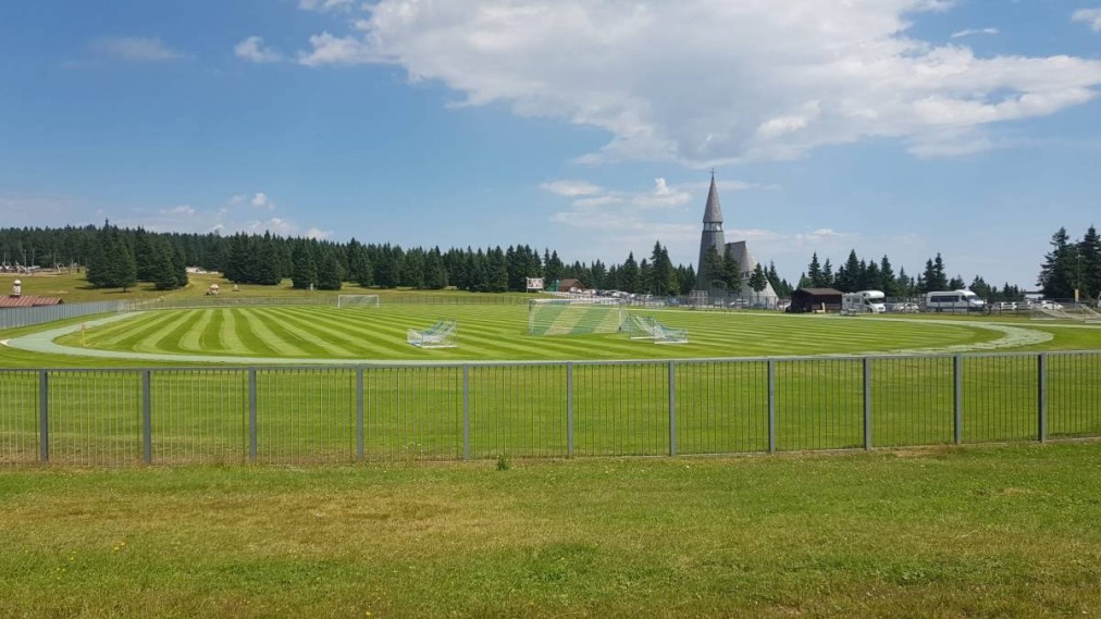 Outdoor football field with a running track and multiple training goals, set against a forest backdrop.
