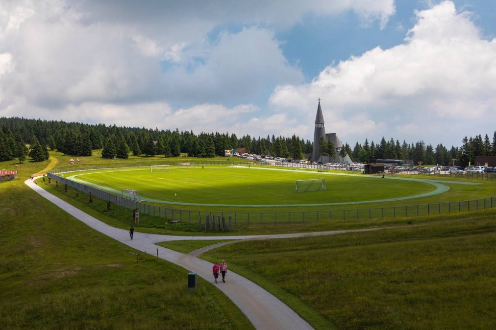 Panoramic view of an outdoor football pitch surrounded by an oval running track, with forested hills nearby.