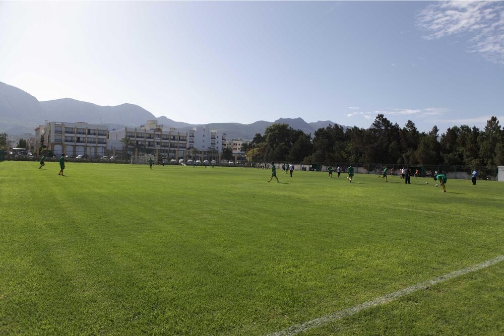 Wide view of a natural-grass football pitch with players training and mountains in the background.