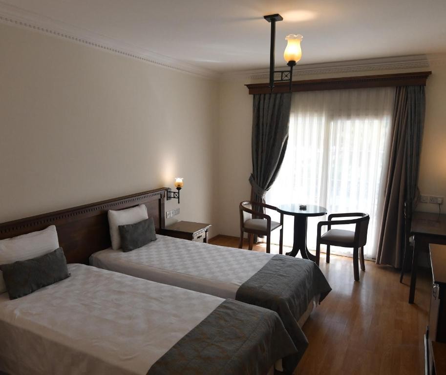 Twin-bed hotel room with small round table and balcony door with curtains.