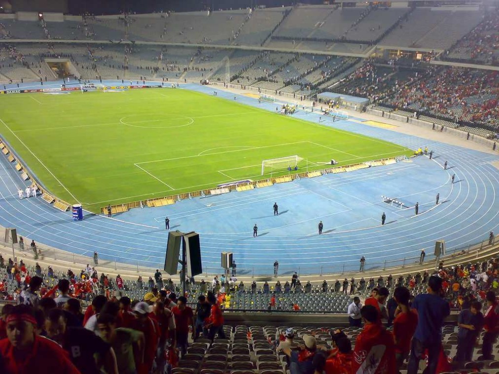 Stadium stands with spectators beside a blue running track and football field.