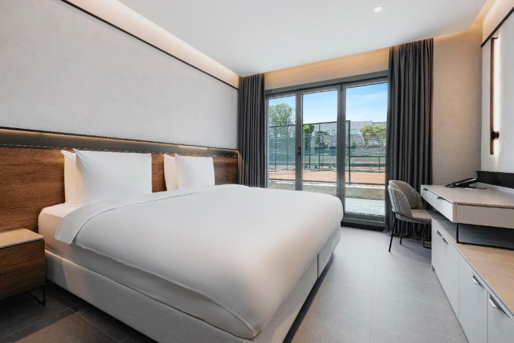 Modern double room with king-size bed, workspace, and balcony doors overlooking the tennis area.