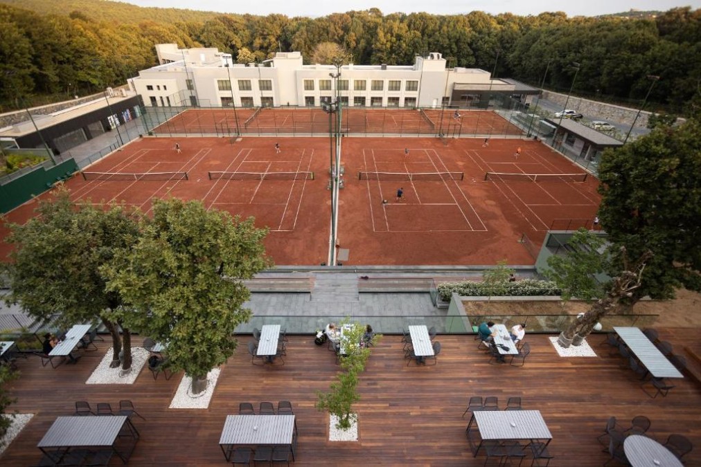 Overhead view of multiple clay tennis courts with terrace seating and surrounding trees.