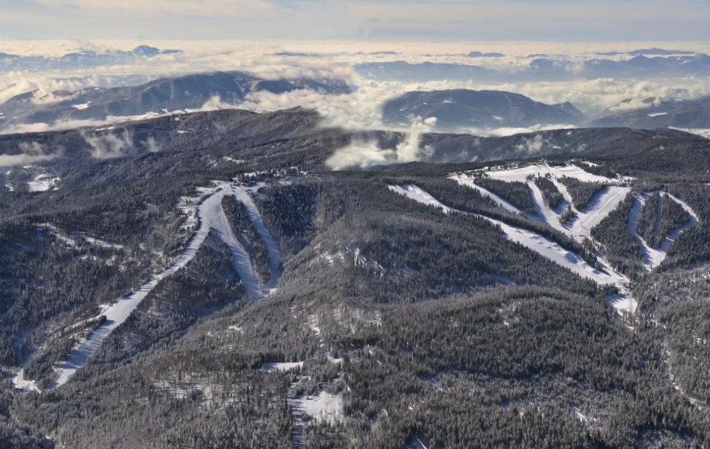 Aerial view of ski slopes and forested mountain ridges above the clouds.
