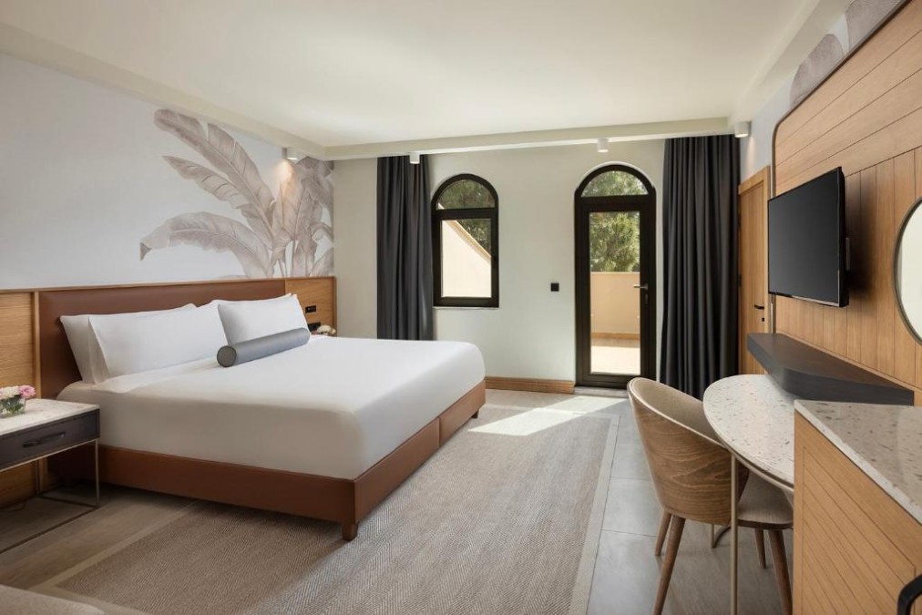 Modern king room with a work desk, wall-mounted TV and balcony door framed by arched windows.