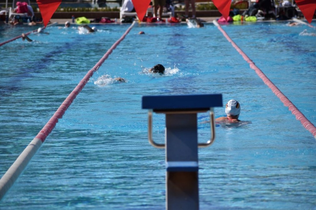 View down an outdoor lap lane with a starting block in the foreground and swimmers in training.