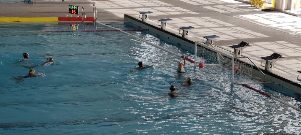 Water polo action near the goal with players defending and attacking.