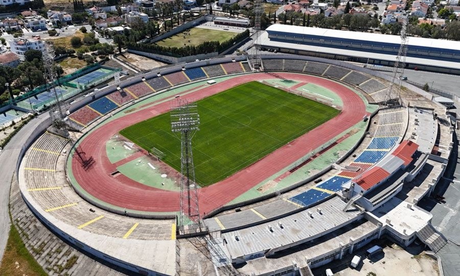 Aerial view of a stadium with a football pitch, running track and adjacent tennis courts.