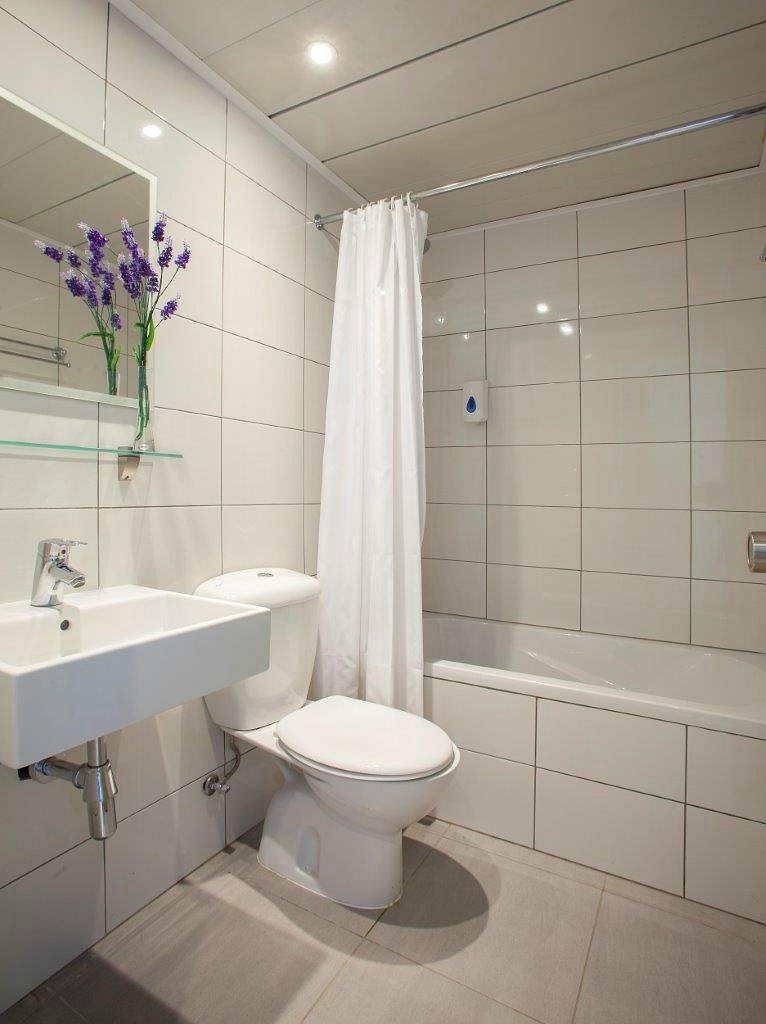 Modern tiled bathroom with sink, toilet, bathtub, shower curtain, and decorative lavender flowers on a glass shelf