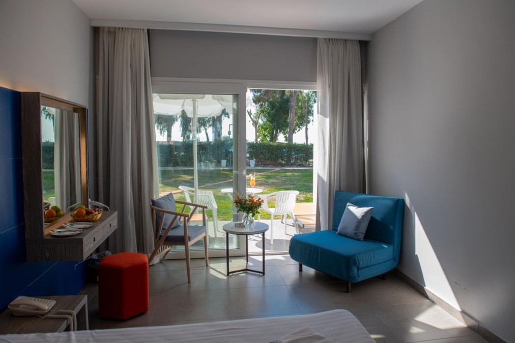 Bright hotel room with blue and gray furniture, a terrace view of a green garden, and a small outdoor seating area