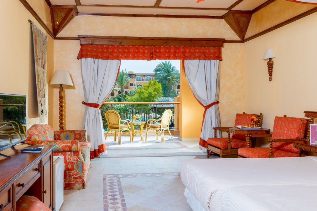 Hotel room with double bed, red decor, and balcony with garden view