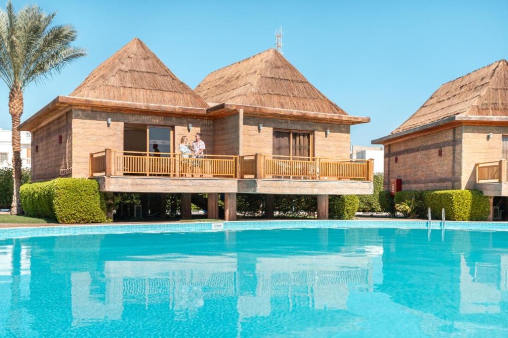 Poolside bungalows on stilts with thatched roofs beside a large outdoor swimming pool.