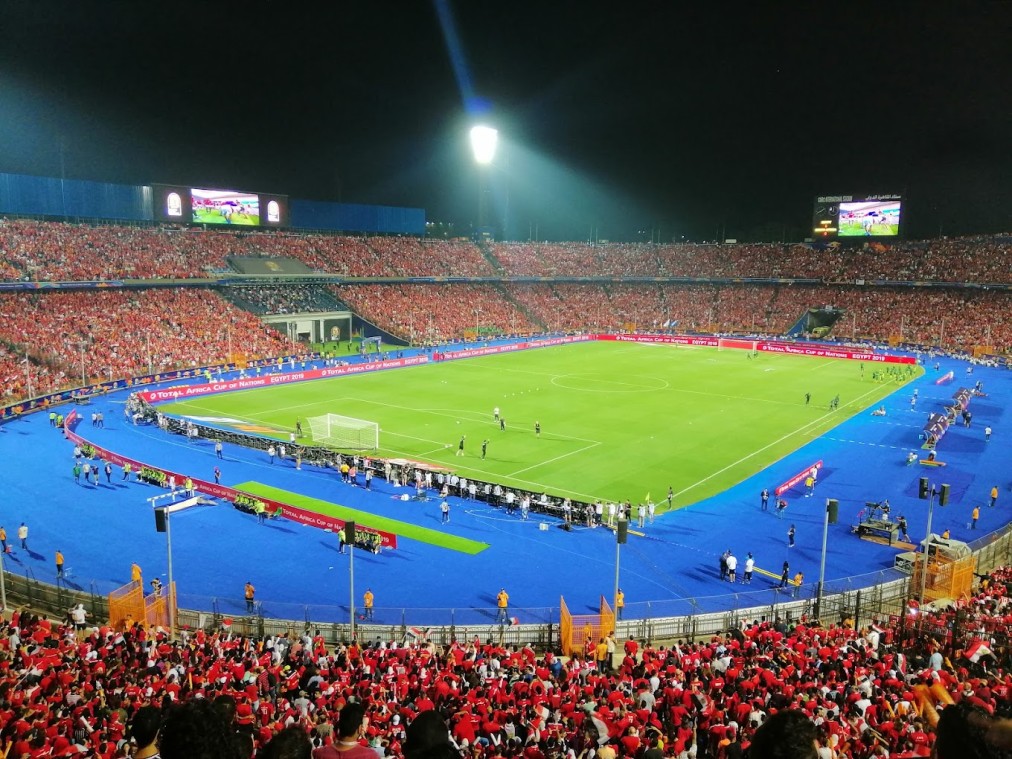 Night match atmosphere in a packed football stadium under floodlights, with a blue track around the pitch.