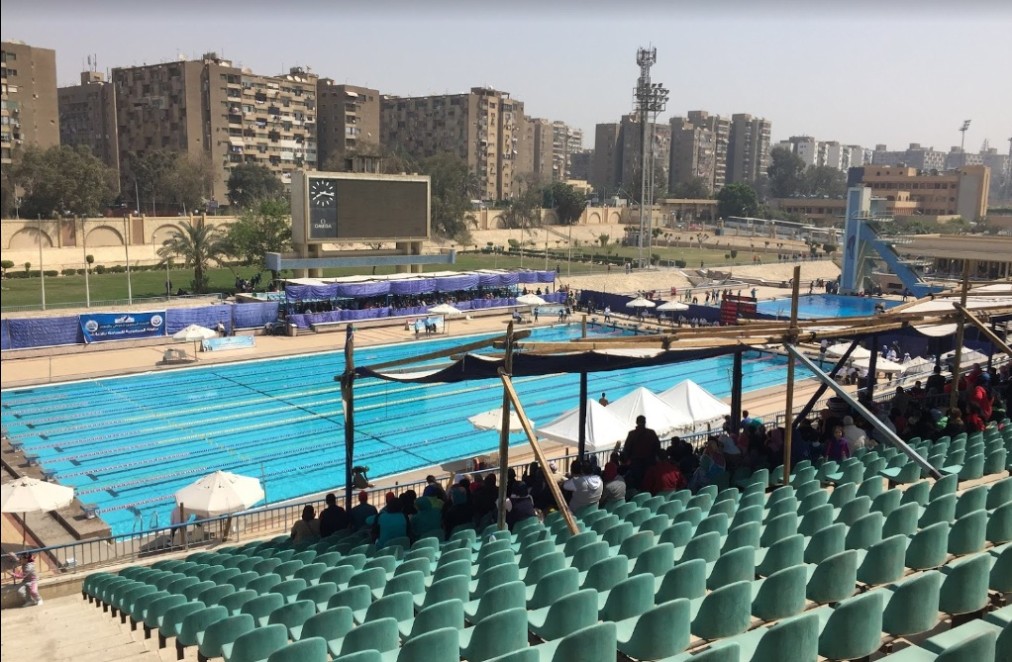 Outdoor 50m pool with lane ropes, spectator stands, and a large scoreboard.