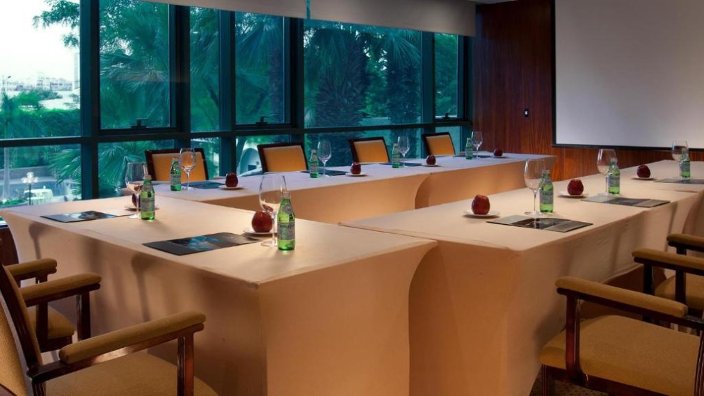 Conference room with U-shaped tables set with water bottles, glasses, and fruit, beside large windows and a screen.