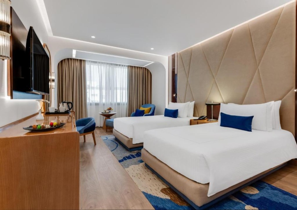 Modern twin room with two beds, desk, wall TV, and armchairs by the window.