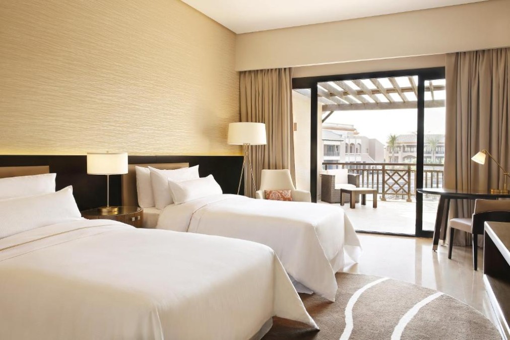 Twin guest room with two beds, work desk, and sliding doors to a balcony overlooking the resort.
