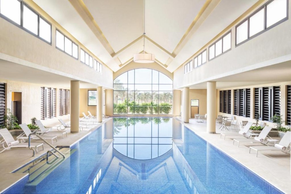 Indoor swimming pool hall with loungers and floor-to-ceiling windows.
