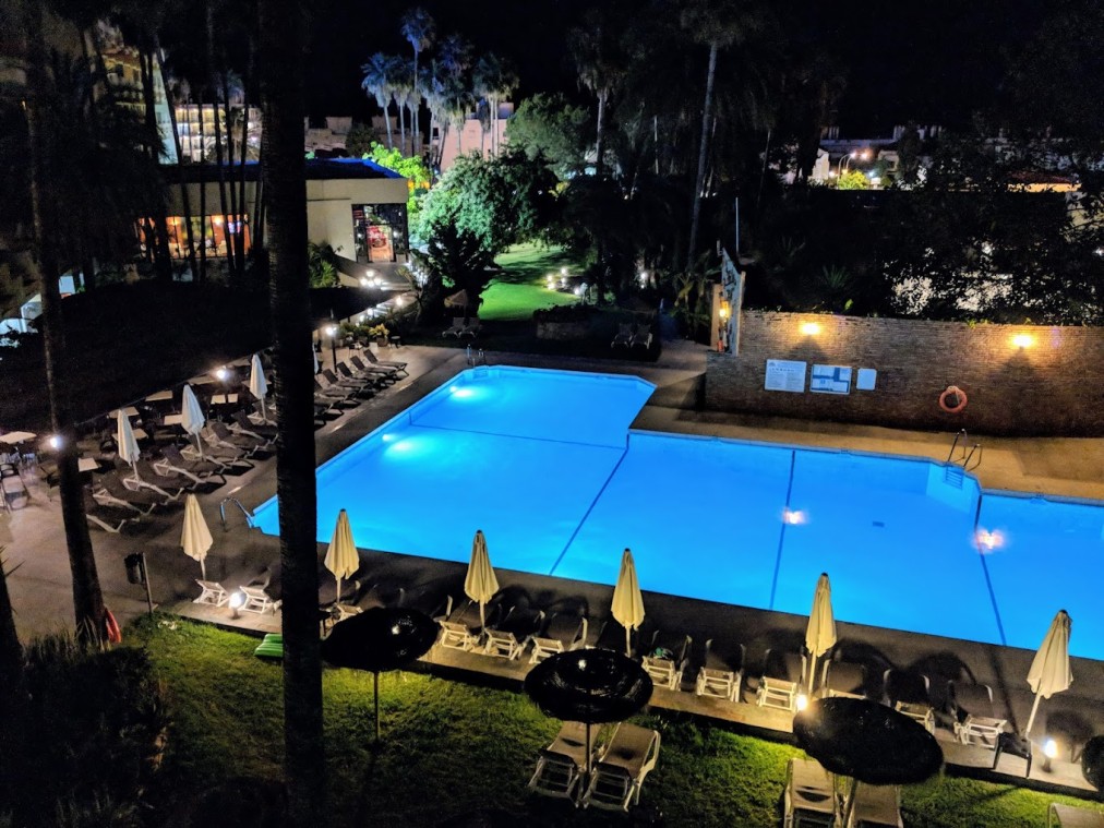 Illuminated outdoor pool with sun loungers and palm trees at night.