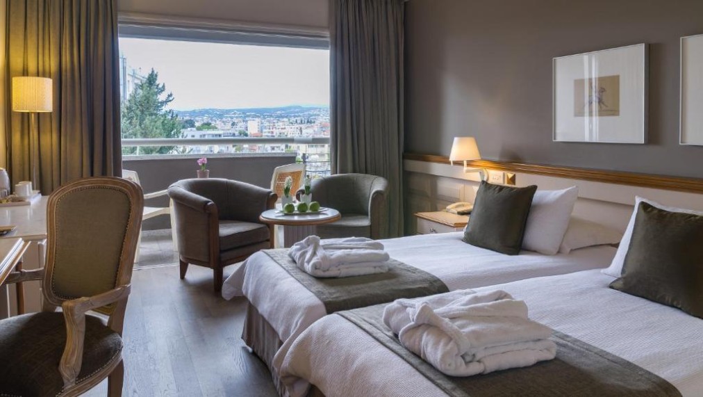 Twin guest room with balcony view, desk and armchairs.