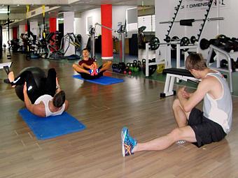 Athleten führen Bodenübungen auf Matten aus, im Hintergrund sind Fitnessgeräte zu sehen