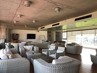 Loungebereich mit Korbmöbeln, weißen Kissen und großen Fenstern mit Blick auf die Sportanlagen