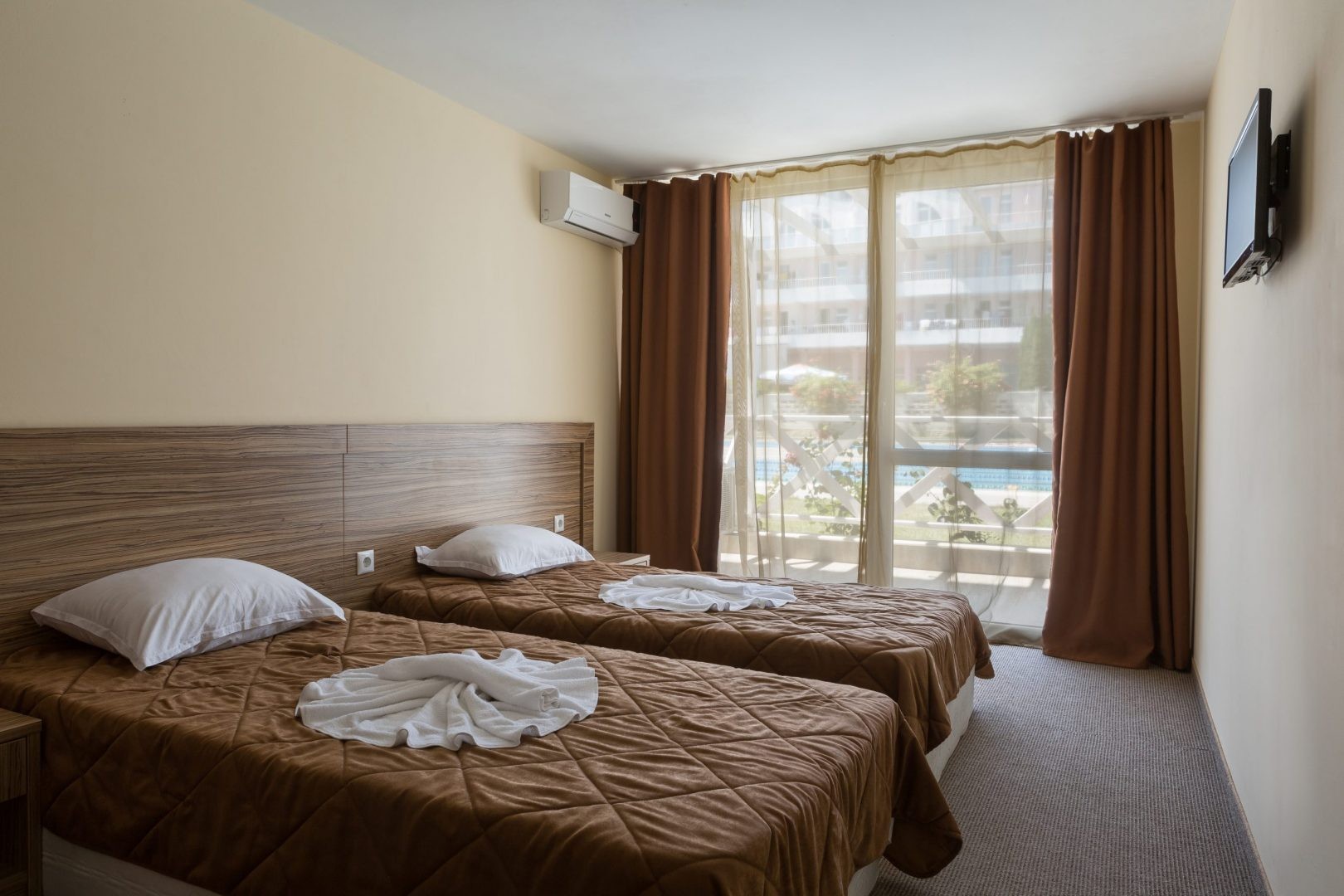 Twin-bed hotel room with brown bedding and balcony view, lodging for athletes during training camp