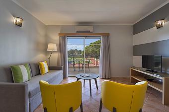 Apartment-Wohnzimmer mit Sofa, zwei Stühlen, TV und Balkonblick auf die Laufbahn.